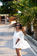 Young beautiful woman in white dress and sunglasses turns around on the road with palms. The concept of joy, ease and freedom during the vacation. The girl is enjoying the rest. Fashion