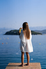 Young beautiful woman in white dress and sunglasses blowing soap bubbles pier with seaview background.. The concept of joy, ease and freedom during the vacation. The girl is enjoying the rest. Back