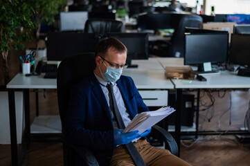 A man in a business suit and medical mask reads a paper report in an empty open space office. Social distance and isolation of employees. Urgent work during quarantine.