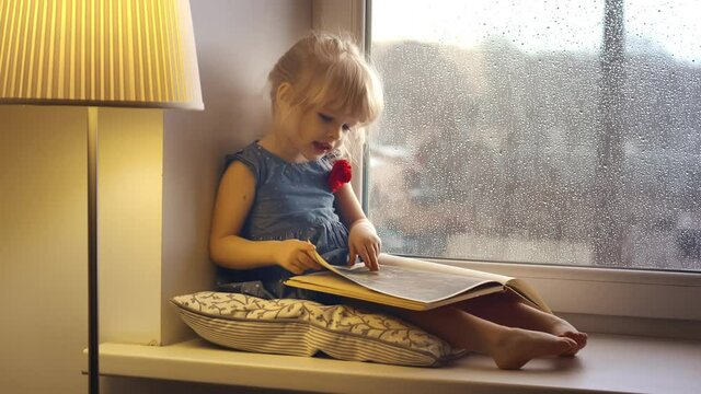 Cute baby girl reading book on window sill at home.