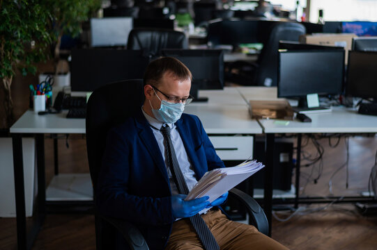 A man in a business suit and medical mask reads a paper report in an empty open space office. Social distance and isolation of employees. Urgent work during quarantine.