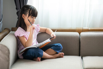 Asian little girl thinking while reading a book on the sofa in the room.