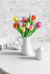 Bouquet of colorful tulips, ceramic white teapot and teacup on the table in the bright kitchen. Cozy home concept