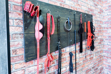 A set of varied leather lashes for sexual pleasures hang on a wooden hanger on a brick wall. Sex equipment in red and black for role-playing games. BDSM room. - obrazy, fototapety, plakaty