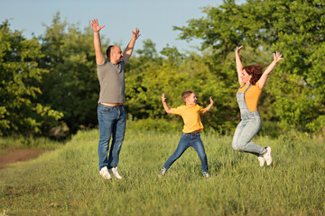 Below is a fun family: mother. father, son in casual clothes having fun and jumping with raised hands in a green field on a summer evening