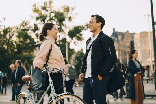 Smiling woman with bicycle looking at male friend while standing in city
