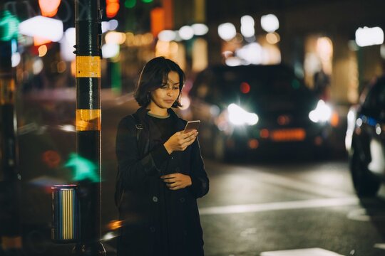 Woman text messaging through smart phone while standing in city at night