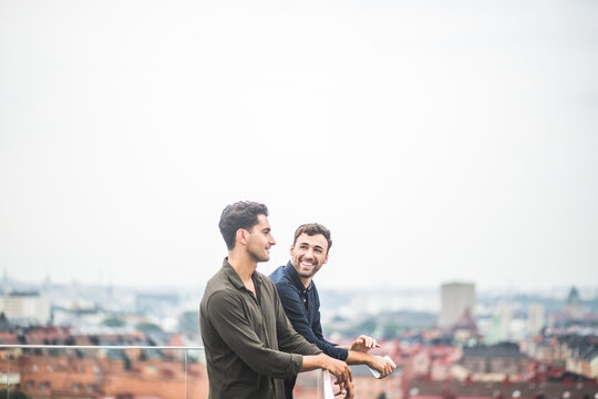 Smiling man looking at male friend while leaning over glass railing on rooftop against sky
