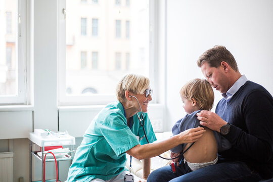 Smiling female doctor examining boy's back with stethoscope while sitting by father in hospital
