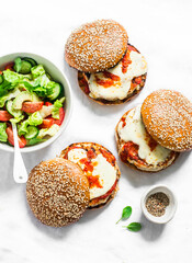 Homemade whole grain bun, grilled chicken, tomato sauce, mozzarella cheese burgers and tomato, cucumber, avocado salad on a light background, top view