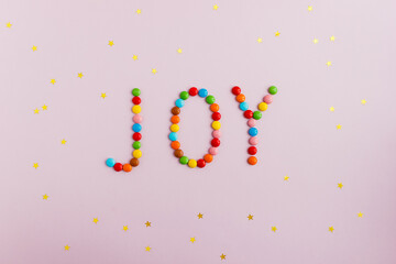 The word Joy composed of multi-colored chocolates on a pink background and decorated with gold star...