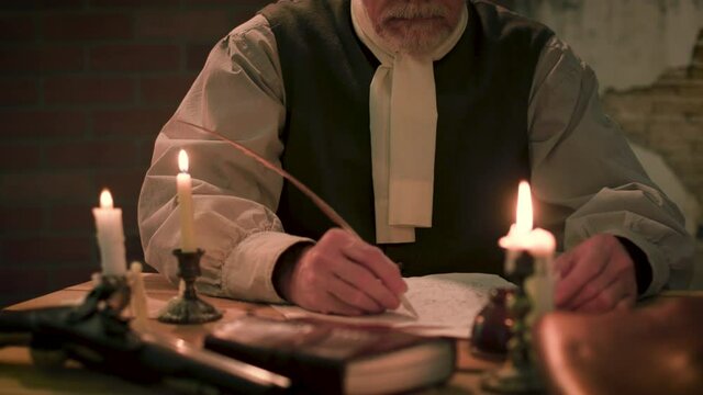Candle lit scene tilts up from a desk with a flintlock pistol laying on it to the face of an American 18th century colonist writing a letter with a plumed quill pen.