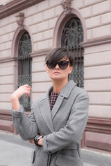 Autumn fashion outdoor. The brunette girl with short hair in fashionable stylish grey coat and sunglasses, posing on the background of building. Street fashion.Autumnal lifestyle