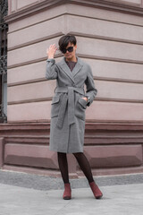 Autumn fashion outdoor. The brunette girl with short hair in fashionable stylish grey coat and sunglasses, posing on the background of building. Street fashion.Autumnal lifestyle