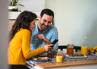 Obraz premium Smiling couple looking in smartphone and having breakfast together stock photo