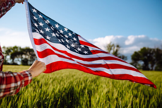 Flag of United States of America flies on the wind. Boy walking with the american flag on the green wheat field celebrating national independence day. 4th of July concept.