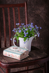 Fresh flowers of forget-me-not in a white kettle instead of a vase and a notebook on an old wooden chair.