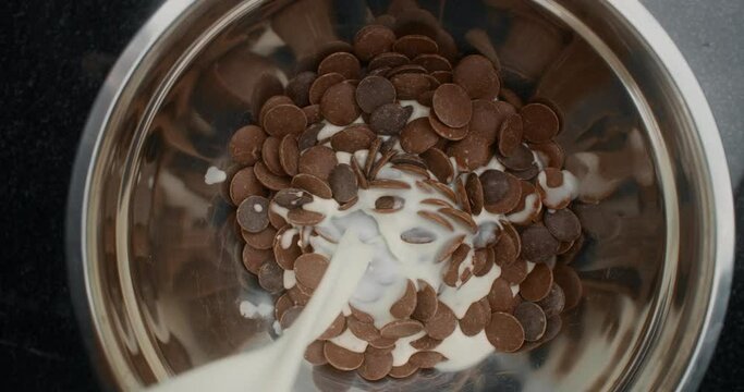 TABLETOP slowmotion shot: chef chocolatier adds hot milk to the metal bowl with shunks of milk and dark chocolate, raw materials for making sweets, cooking the desserts, making chocolate bars, 4k 120p