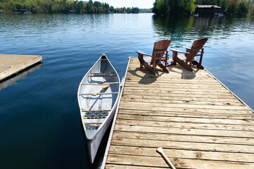 Two Muskoka chairs sitting on a wood dock facing a lake. Across the calm water is a brown cottage nestled among green trees. A canoe is tied to the dock, life jackets and canoe paddles are visible.