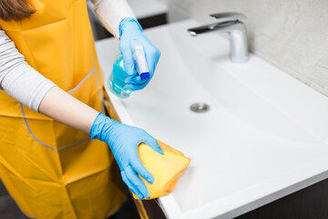 Close up of female hands in rubber gloves cleaning bathroom basin edge with sprayer disinfectant and microfiber cloth. Hygiene antibacterial concept.
