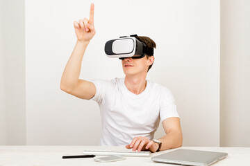 Male using virtual reality headset interacts with augmented things orienting in three dimensional space while sitting on his desk with keyboard and laptop on white background.