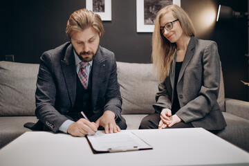 Group of two business partners in formal clothing sitting on grey couch and signing documents. Beautiful woman and bearded man successfully ending meeting at modern office.