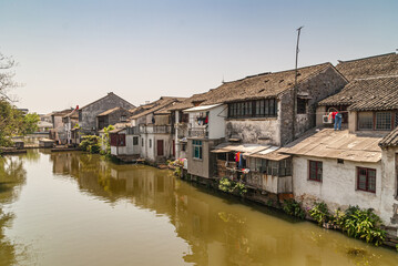 Fototapeta na wymiar Tongli, JIangsu, China - May 3, 2010: Line of dark roofed dirty poor white houses reflected in greenish water of canal under light blue sky. Green foliage and laundry add color.