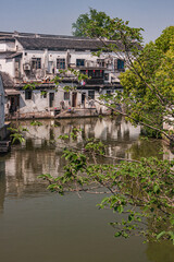 Fototapeta na wymiar Tongli, JIangsu, China - May 3, 2010: Line of dark roofed white houses reflected in brown water of canal under light blue sky. Green foliage and red lanterns add color.