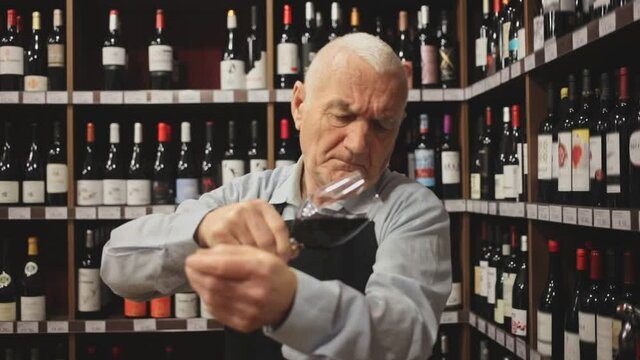 Confident elderly male winemaker holding glass of red wine, checking it in wine store