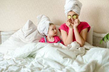 Obraz na płótnie Canvas two cute sisters sitting on bed in white towel,applying cucumber slices to her eyes, Baby girls have a funny face.Morning facial, cosmetology. Adult mimicry. Teen first makeup.