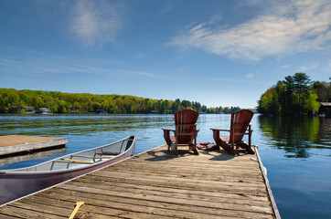 Two Muskoka chairs sitting on a wood dock facing a lake. A canoe is tied to the dock. Life jackets...