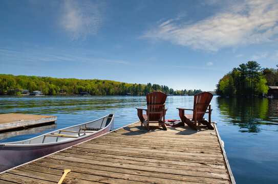 Two Muskoka chairs sitting on a wood dock facing a lake. A canoe is tied to the dock. Life jackets are visible near the chairs.