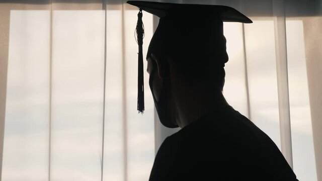 The rear view of the graduate in a cap and robe looks out the window. He's wearing a protective mask during the epidemic.