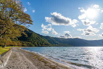 Beautiful autumn foliage scenery landscapes of Lake Towada in sunny day. View from lakeside, clear sky, blue water and white cloud in the background. Towada Hachimantai National Park, Aomori, Japan
