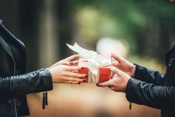 Cropped shot of hands and giftbox, man gives his girlfriend a present tied with white ribbon over blurred autumn background.