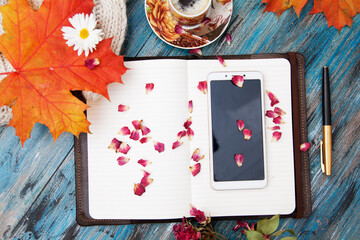 Autumn layout, notebook and pen, smartphone, cup of coffee, orange leaves, dry rose petals, knitted sweater. Cozy workplace, top view