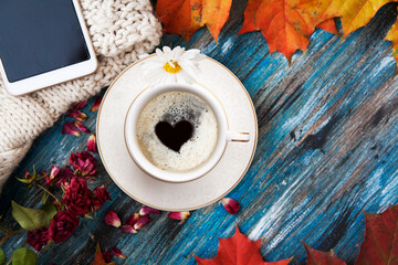 Autumn layout, a cup of coffee with a heart inside the foam, orange leaves, dry roses and petals, a knitted sweater, a smartphone with an free space for text