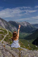 Trip to Norway. Young woman in striped dress feels freedom alone over a cliff and looking far at Trollstigen road and green valley with high rocky mountains on background in bright summer day