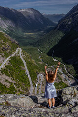Trip to Norway. Young woman in striped dress feels freedom alone over a cliff and looking down at Trollstigen road and green valley with high rocky mountains on background in bright summer day
