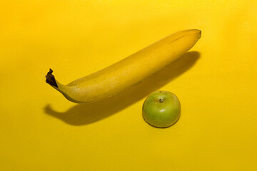 A yellow banana flies levitating over a yellow background casting a shadow next to a fresh green apple