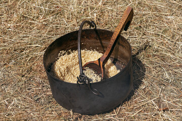 Natural cereal porridge in a smoked metal forged pot with a wooden spoon stands on yellow grass
