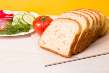 bread for toast with slices of vegetables lies on a wooden cutting board, in the background ingredients for a sandwich, vegetables and sausage are cut on a plate, yellow background
