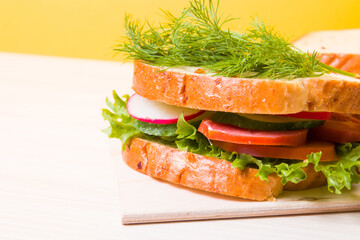 big sandwich of vegetables and diet sausage on a wooden cutting board, yellow background, bread and vegetables in the background, healthy breakfast concept