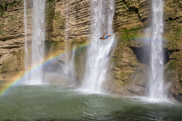 Cliff diver with waterfall in background jumps in the water against a rainbow.