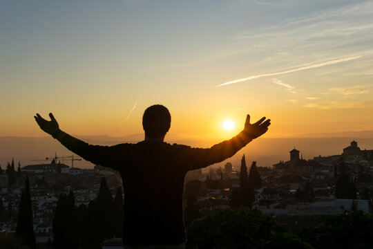 Picture of a man shot from behind looking at the sunset over Granada with his hands up in the air. 