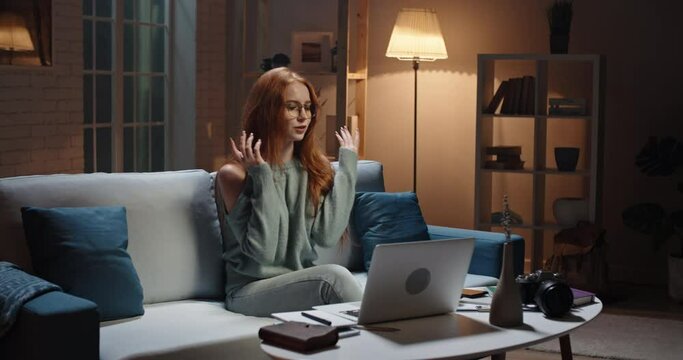 Pretty caucasian redhead girl sitting on coach looking at laptop, having a videochat or online tutorial, recording an internet vlog, cute model looking at webcamera and talking 4k footage