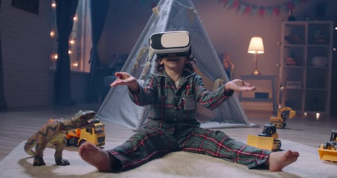 Little boy with curly hair wearing pyjamas is playing video games before bedtime, using virtual reality headset and experiencing new world 4k footage