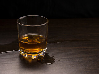 A glass with whiskey and spilled liquid on a dark wood table on a dark background.