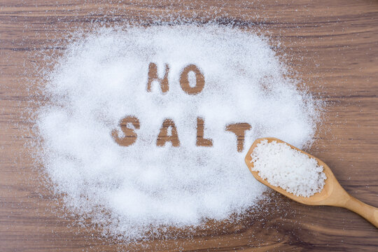 White granulated natural sea salt in wooden scoop and words" no salt " letters written in salt grains on wood table background. Unhealthy food concept. Overhead view. Flat lay.