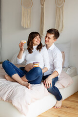 Fototapeta na wymiar Pair of future parents laying on bed eating donuts. Pregnant woman hugging her huband in bedroom at home. Family at Cozy home. Romantic parents waiting for baby. Casual clothes outfit. Rest together